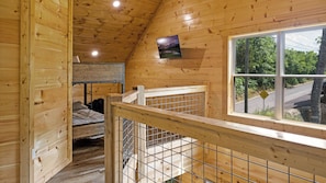 Interior de una cabaña de madera con literas, un televisor montado, una ventana con vista a una carretera y una puerta que conduce a otra habitación.