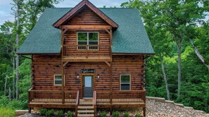A two-story log cabin with a balcony, a welcome sign, and a front porch.