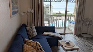 Un salon avec un canapé bleu foncé, une petite table surmontée d’un set de table en fibres tressées, une lampe sur pied et une vue sur une piscine et l’océan, visible à travers une porte-fenêtre coulissante.
