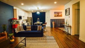A modern living room with a dark blue sofa, wooden flooring, and a kitchen in the background. - 4 BR , Large Yard with HOT TUB, Fire pit,Basket ball hoop (Dallas)