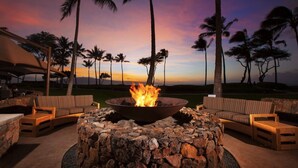 A fire pit surrounded by stone seating, palm trees, and a sunset view.