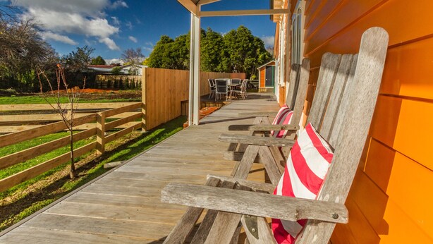 A covered outdoor seating area with wooden benches and a table, a wooden fence, and a building with orange siding in the background. - The Lazy Carrot - Ohakune Holiday Home (Ohakune)