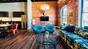 A modern interior with a brick wall, a flat-screen TV, a round table with a plant, two blue chairs, a sofa with floral upholstery, and a dining area with a wooden table and chairs. - One Of A Kind Huge Loft Minutes From Downtown (Detroit)