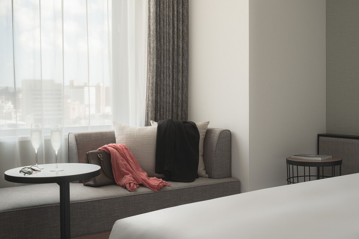 A hotel room with a sofa, a small round table, a chair, and a view of the city through the window.