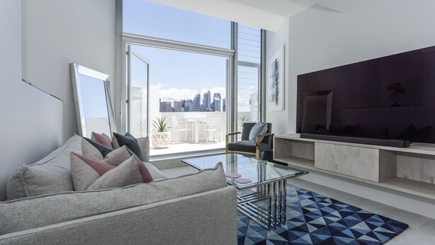 Ein modernes Wohnzimmer mit einem großen Fernseher, einer Couch, einem Glastisch und Blick auf die Stadt durch große Fenster.