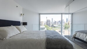 Ein modernes Schlafzimmer mit einem großen Bett, einem zusätzlichen Einzelbett, einem Blick auf die Stadt durch große Fenster und einer städtischen Skyline im Hintergrund.