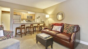 A living room with a brown leather sofa, a coffee table, and a kitchen area with wooden cabinets and a microwave. - Main Street Snowcave walkable to Quicksilver (Breckenridge)