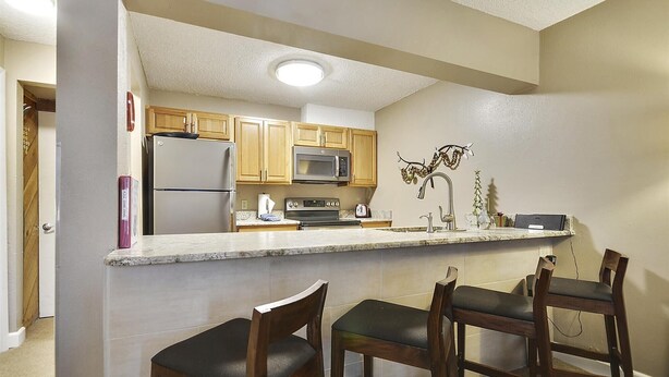 A kitchen with wooden cabinets, a stainless steel refrigerator, a microwave, and a stove. There are three bar stools in front of the counter. - Main Street Snowcave walkable to Quicksilver (Breckenridge)