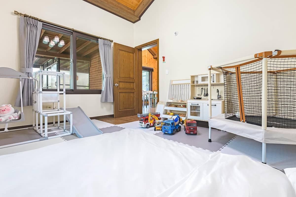 Ein Zimmer mit Etagenbetten, einer Spielecke mit Spielzeug, einem Fenster mit Vorhängen und einer Tür zu einem anderen Raum.