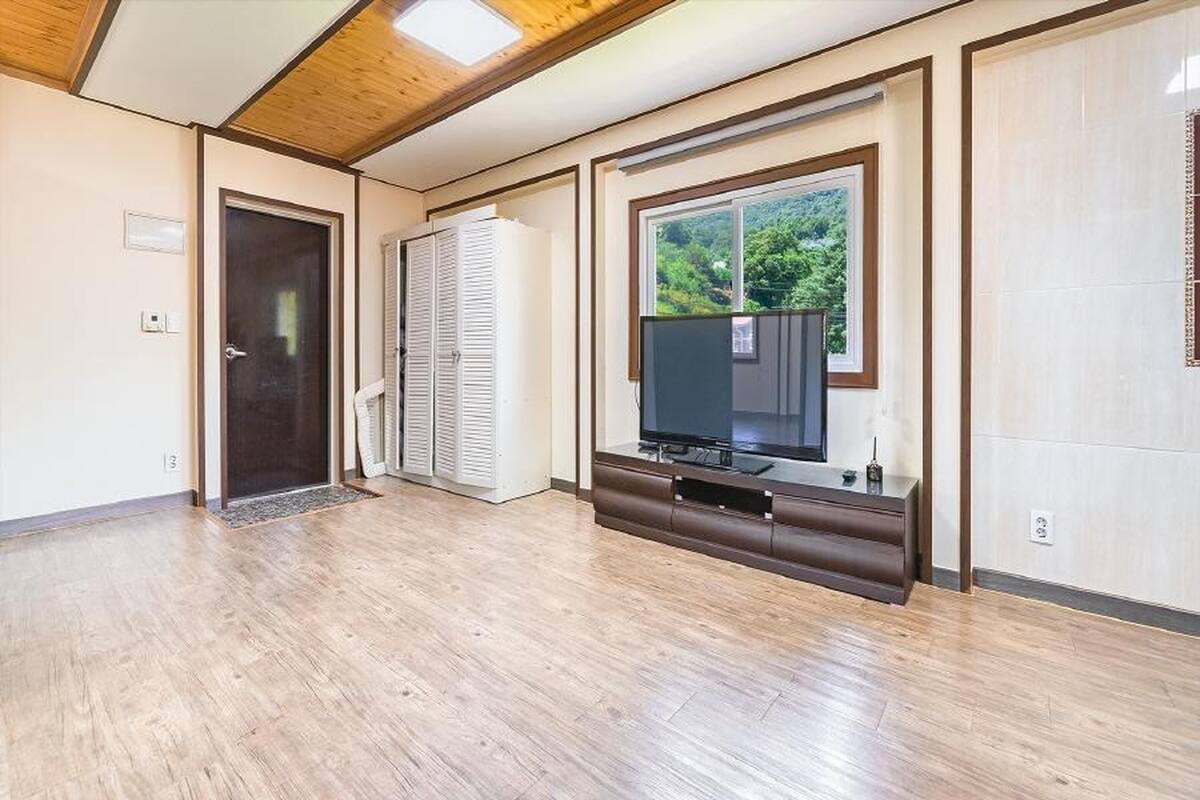 Ein Zimmer mit Holzboden, einem großen Fenster mit Blick auf Grünflächen, einer geschlossenen Tür und einem Fernseher auf einem Holztisch.