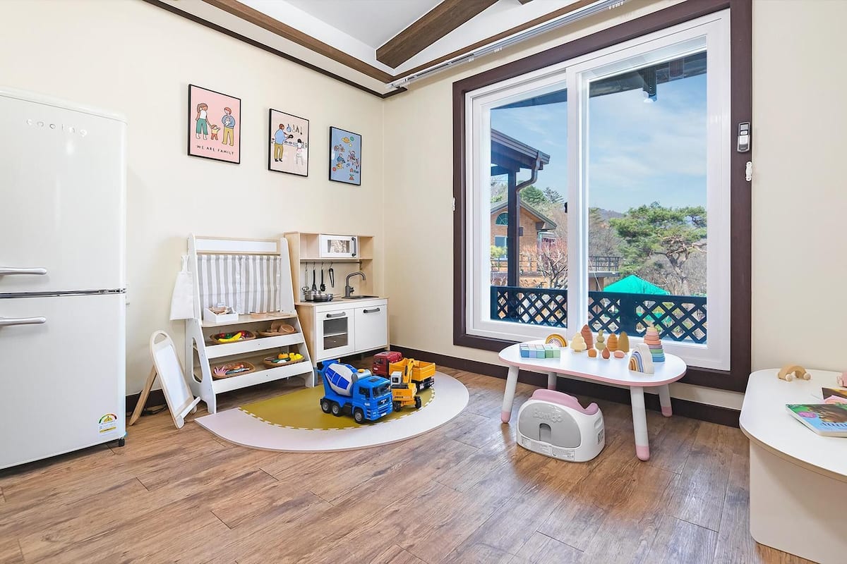 Ein Kinderzimmer mit einem weißen Kühlschrank, einer Spielküche, Spielzeug und einem großen Fenster.