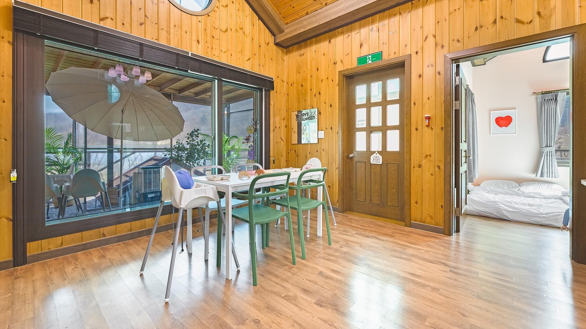 A room with a wooden interior, a dining table with chairs, a large window with an umbrella, and a door with a sign.