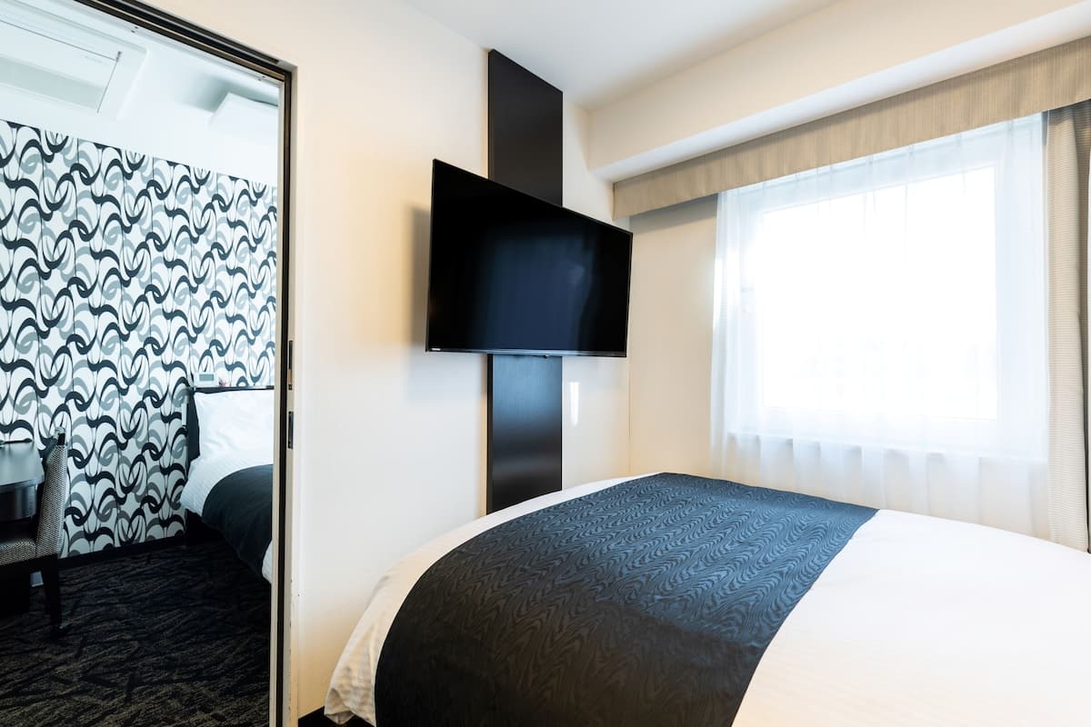 A hotel room with a bed, a flat-screen TV mounted on the wall, a large window with curtains, and a patterned wall.