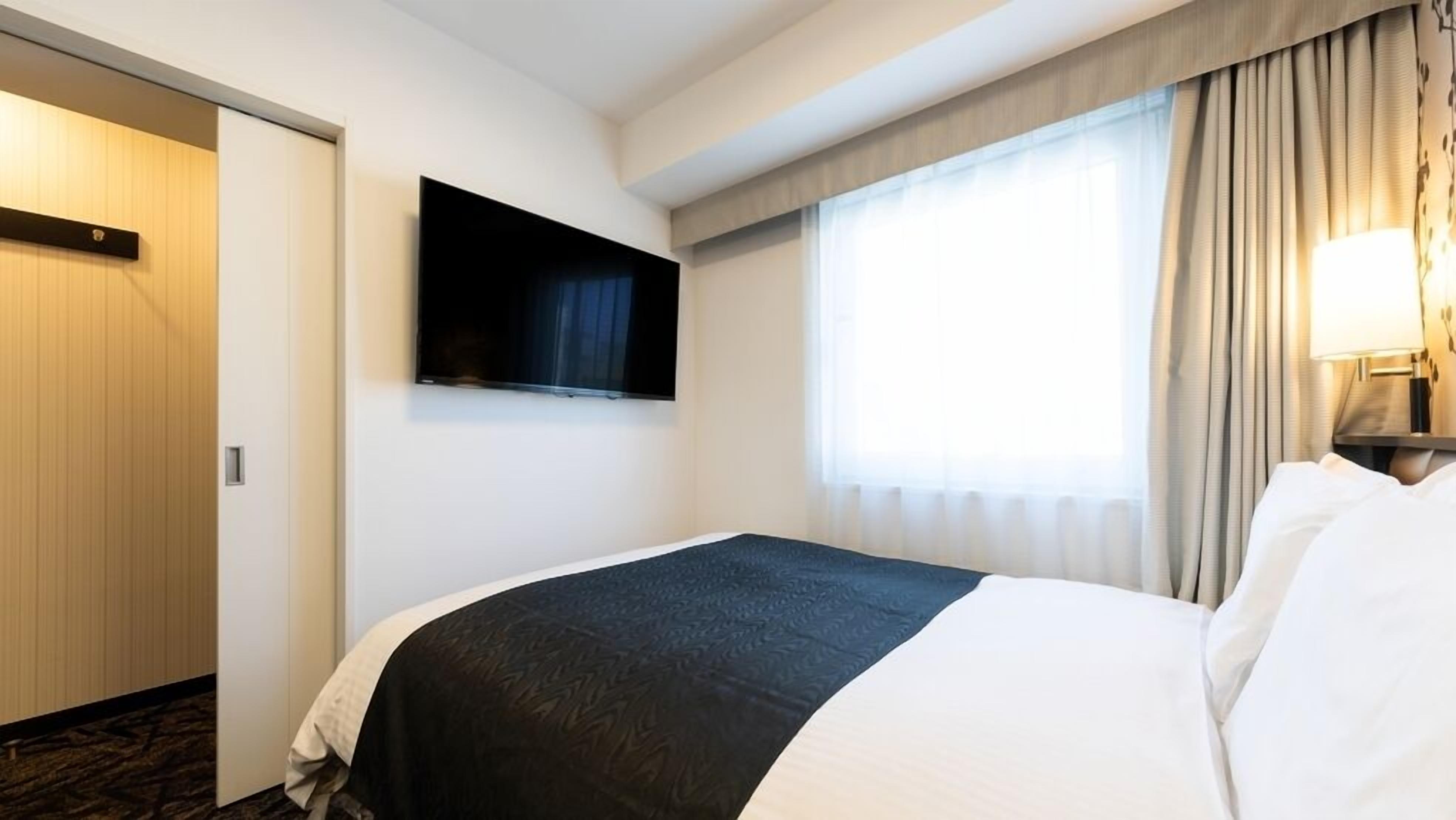 A hotel room with a bed, a television mounted on the wall, a window with curtains, and a lamp on the nightstand.