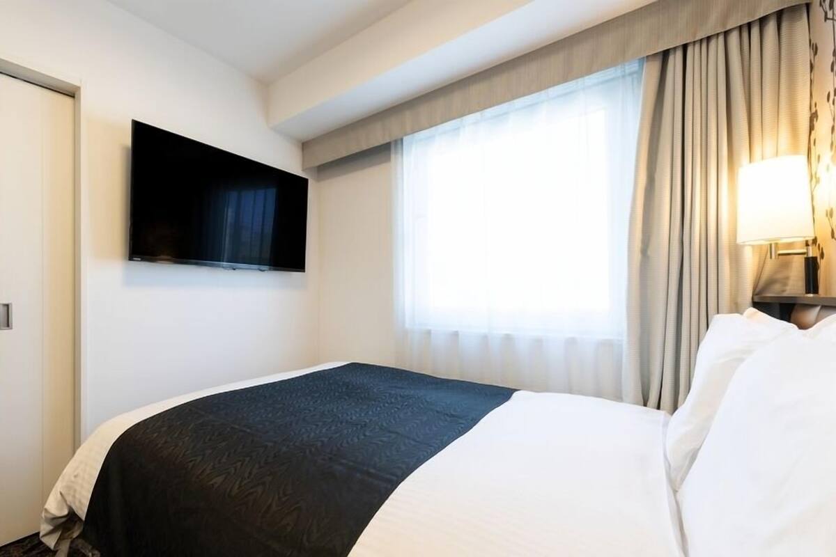 A hotel room with a bed, a television mounted on the wall, a window with curtains, and a lamp on the nightstand.