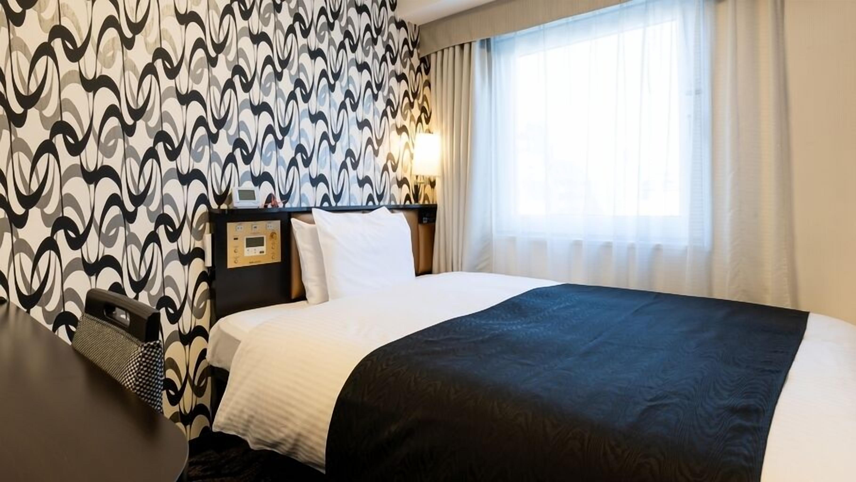 A hotel room with a bed, a desk, a nightstand, a lamp, and a window with curtains.