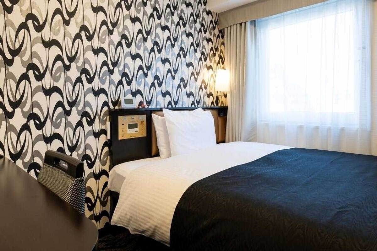 A hotel room with a bed, a desk, a nightstand, a lamp, and a window with curtains.