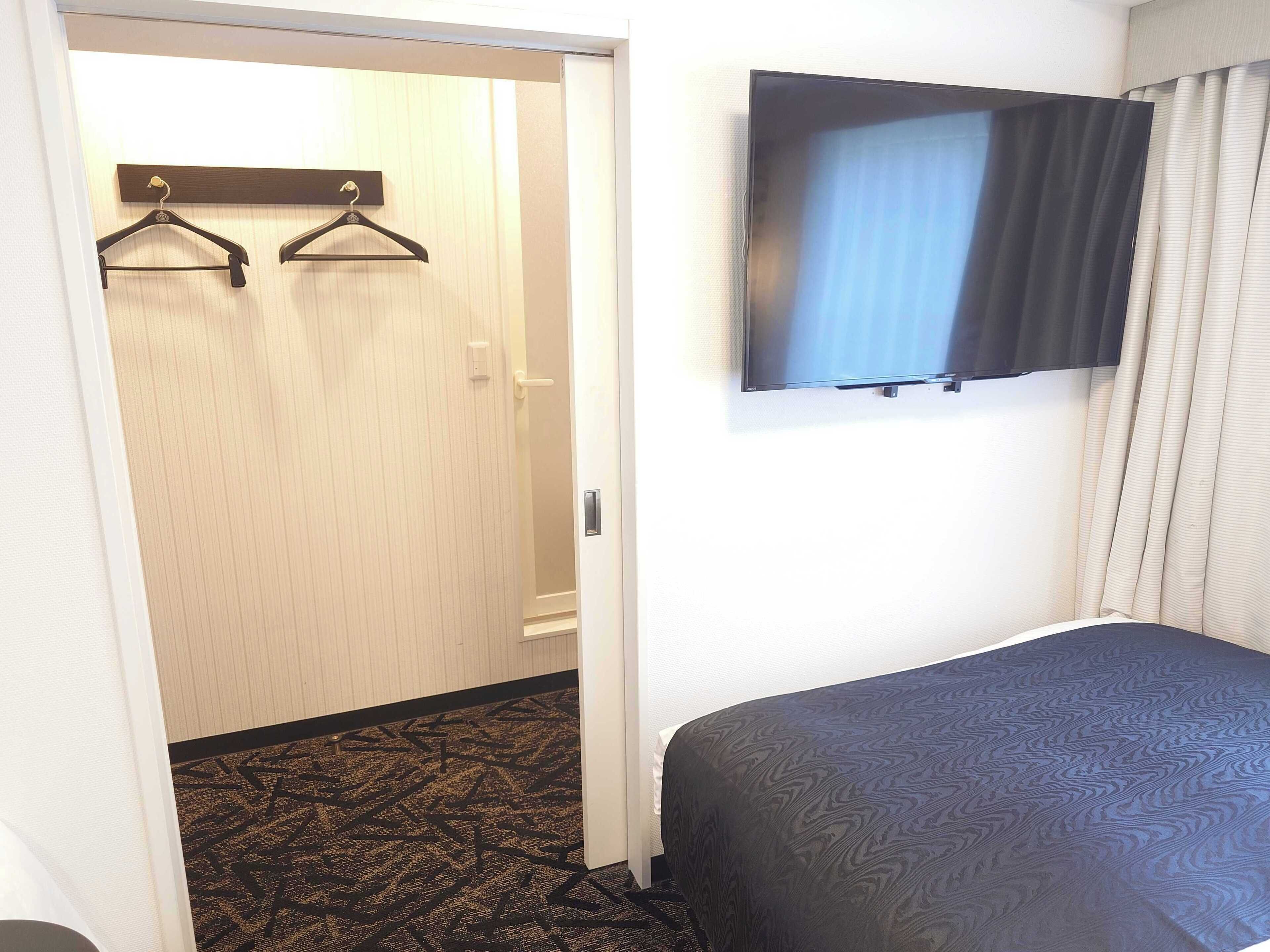 A hotel room with a bed, a television mounted on the wall, a door leading to a hallway, and a coat rack with hangers.