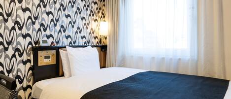 A hotel room with a large bed, a control panel, a bedside table, and a window with sheer curtains.
