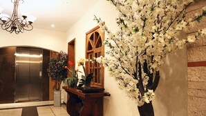 A lobby with a wooden reception desk, a large artificial tree, an elevator, and a chandelier.