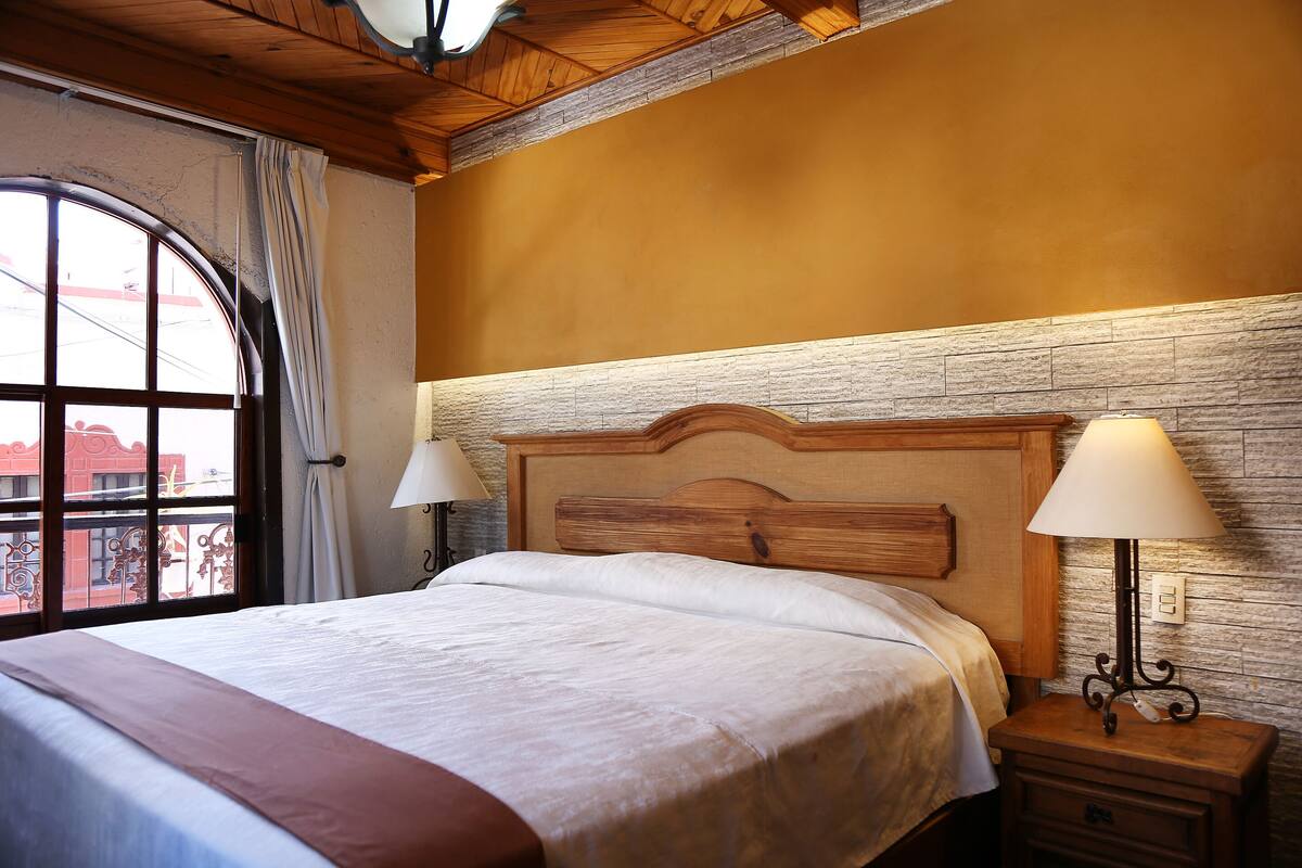 A hotel room with a large bed, wooden headboard, two bedside lamps, a nightstand, and a view through a window with curtains.