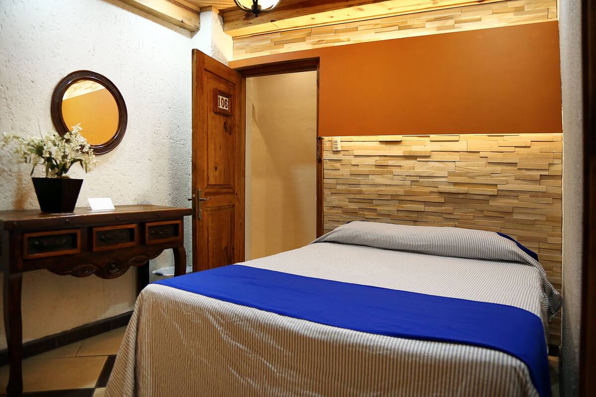A hotel room with a bed, a wooden dresser, a mirror, and a potted plant.