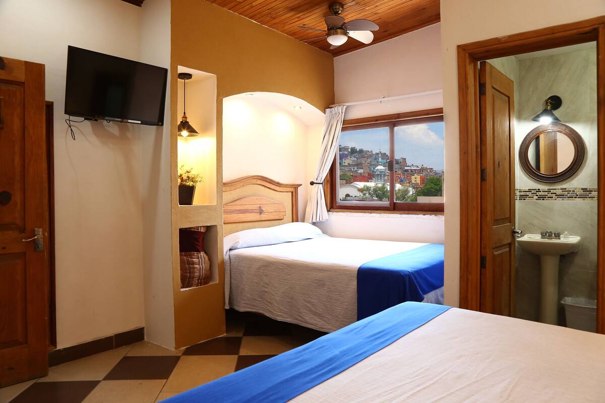 A hotel room with two beds, a television, a window with a view of the city, and a bathroom with a sink and mirror.