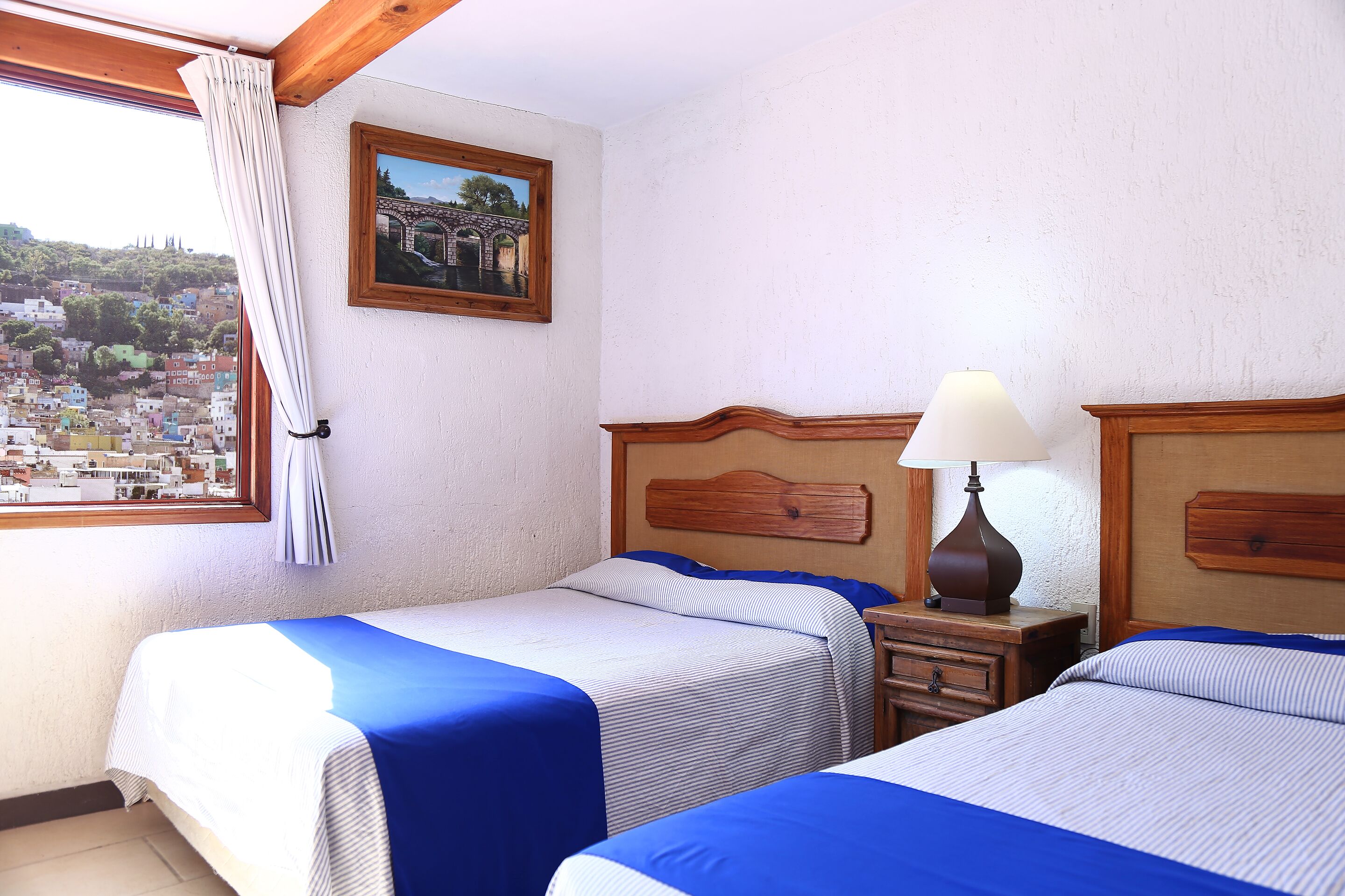 Hotelkamer met twee bedden, een houten hoofdeinde, een nachtkastje met lamp, een raam met gordijnen en uitzicht op de stad.
