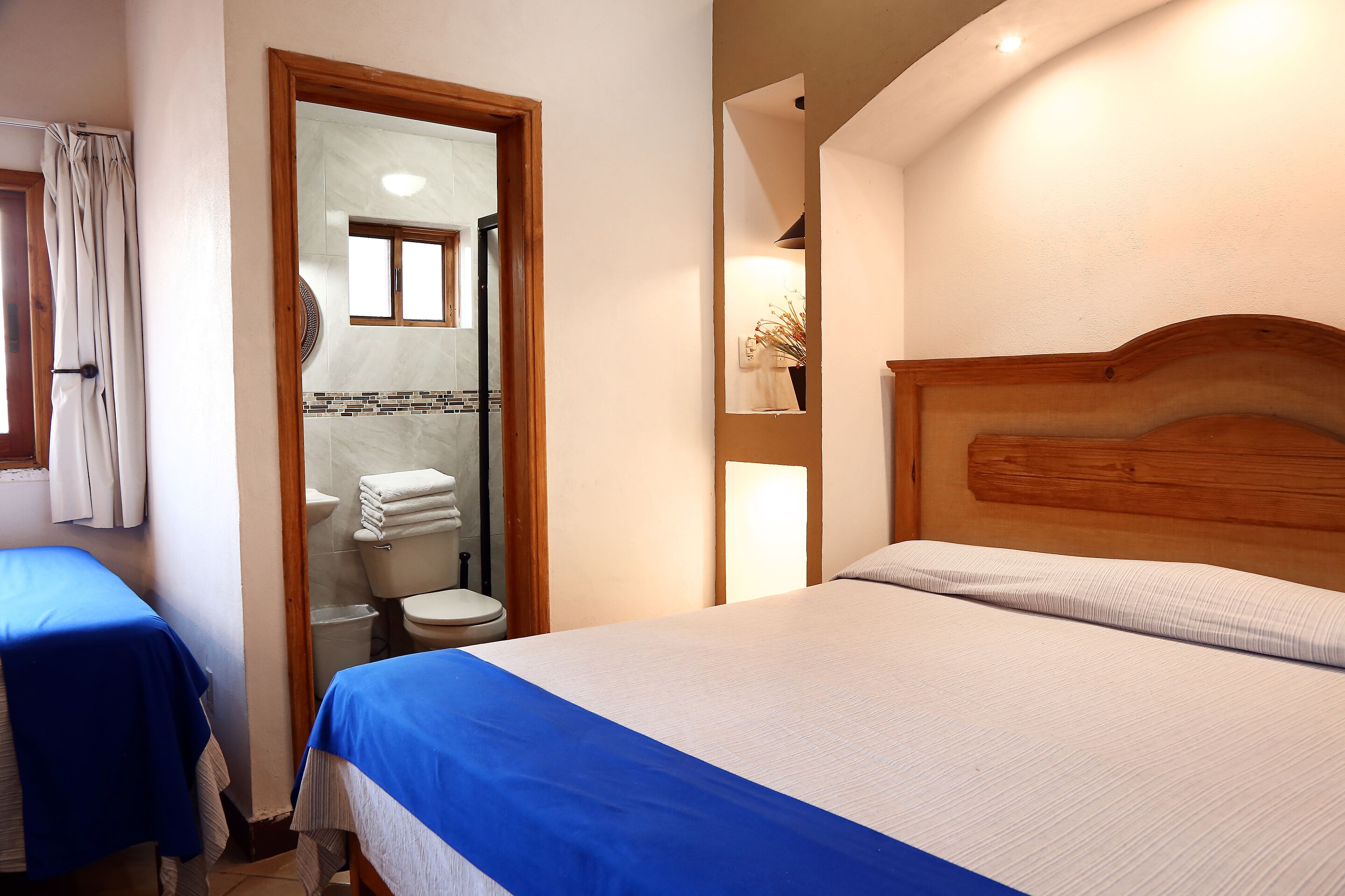 Quarto de hotel com cama, banheiro com vaso sanitário e chuveiro, e janela com cortinas.