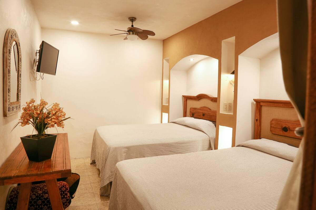 A hotel room with two beds, a desk with a potted plant, a ceiling fan, and a TV mounted on the wall.
