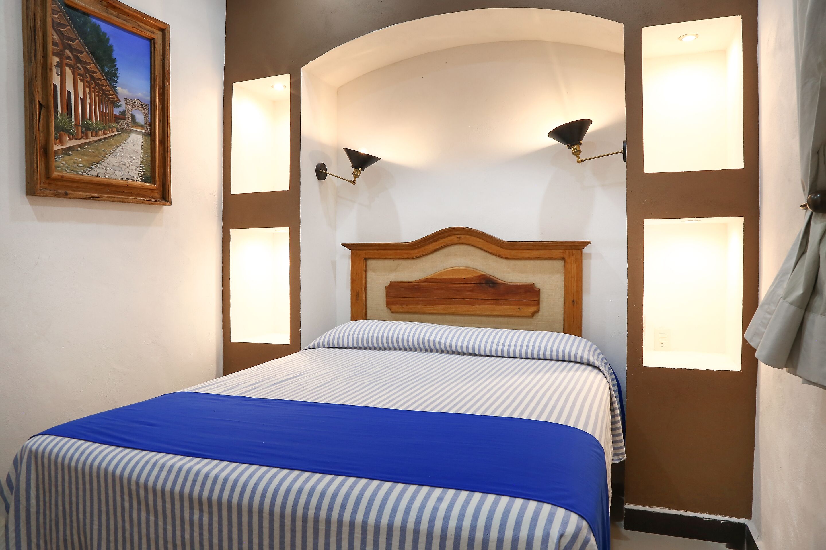 Una cama impecable con ropa de cama a rayas y un detalle azul, un cabecero de madera y lámparas fijadas a la pared.