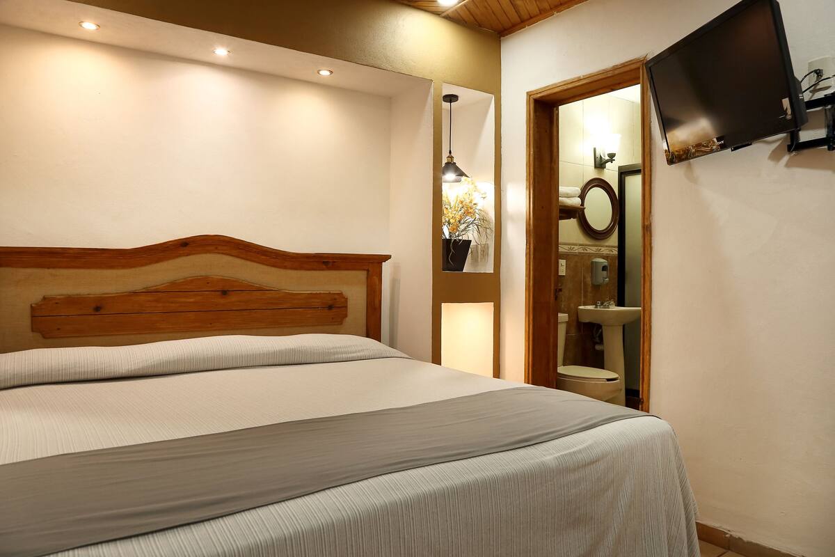 A bedroom with a large bed, a wooden headboard, a bedside lamp, a TV mounted on the wall, and a bathroom visible through an open door.