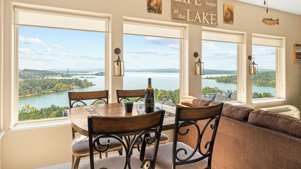 A dining area with a lake view, a ceiling fan, and a sign that reads "Life is better at the lake." - Table Rock View Penthouse with Sun Room, Luxury Decor, Free Tickets & Prime Location (Branson)