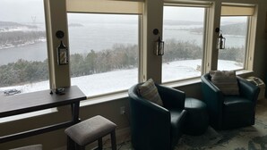 A cozy room with large windows overlooking a snowy landscape and a lake. There are two teal armchairs and a small table with a bench. - Table Rock View Penthouse with Sun Room, Luxury Decor, Free Tickets & Prime Location (Branson)