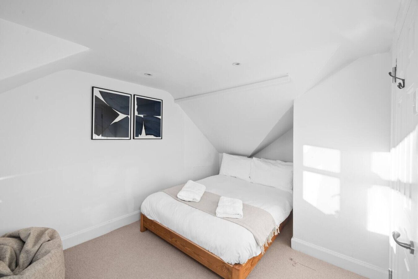 Un dormitorio con techo inclinado, estructura de cama de madera, ropa de cama blanca y dos obras de arte abstracto en la pared.