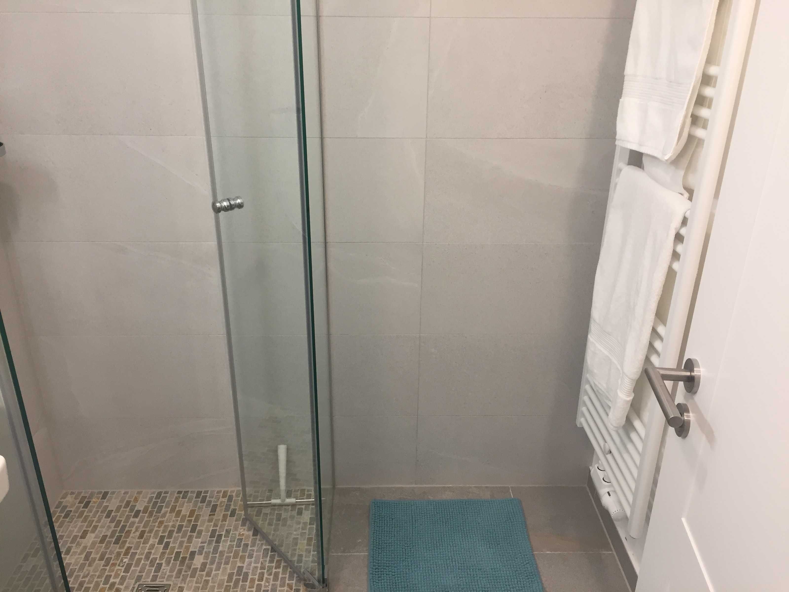 Une douche fermée par des parois de verre, un sol carrelé, un porte-serviettes avec des serviettes blanches et un tapis bleu.