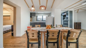 A modern kitchen with a dining area. The kitchen has a large window, a refrigerator, and a sink. The dining area has a wooden table with four chairs. There is a small plant on the table.