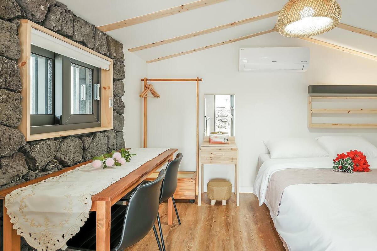 Ein gemütliches Schlafzimmer mit einem Bett, einem Schreibtisch und einem Stuhl. Eine Steinwand, Holzbalken und eine hängende Leuchte sind zu sehen.