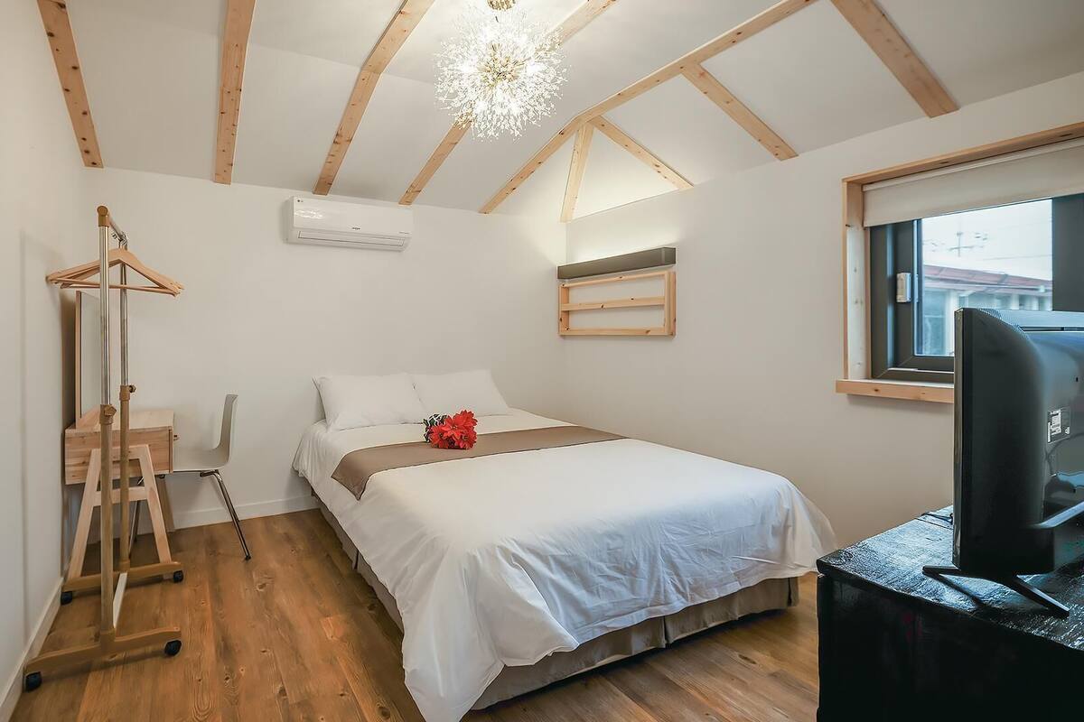 Ein Schlafzimmer mit einem Bett, einem Schreibtisch, einem Stuhl, Holzboden und einer Decke mit sichtbaren Balken, sowie einem Fenster.