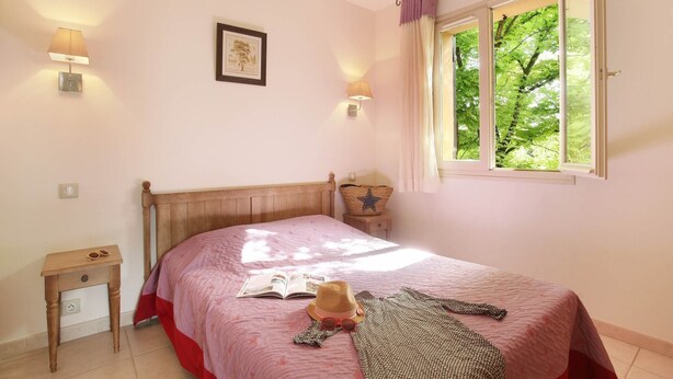 Een slaapkamer met een bed, nachtkastje, lamp, raam en een lijst met afbeelding aan de muur.