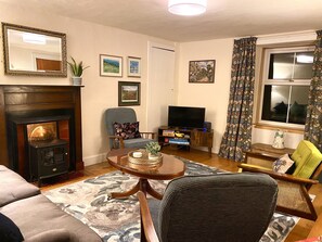 A living room with a fireplace, a television, and a window with curtains. - Wee Silverburn (Isle of Arran)