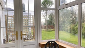 A room with a checkered floor, a black chair, a wooden bench, and a view of a garden through large windows.