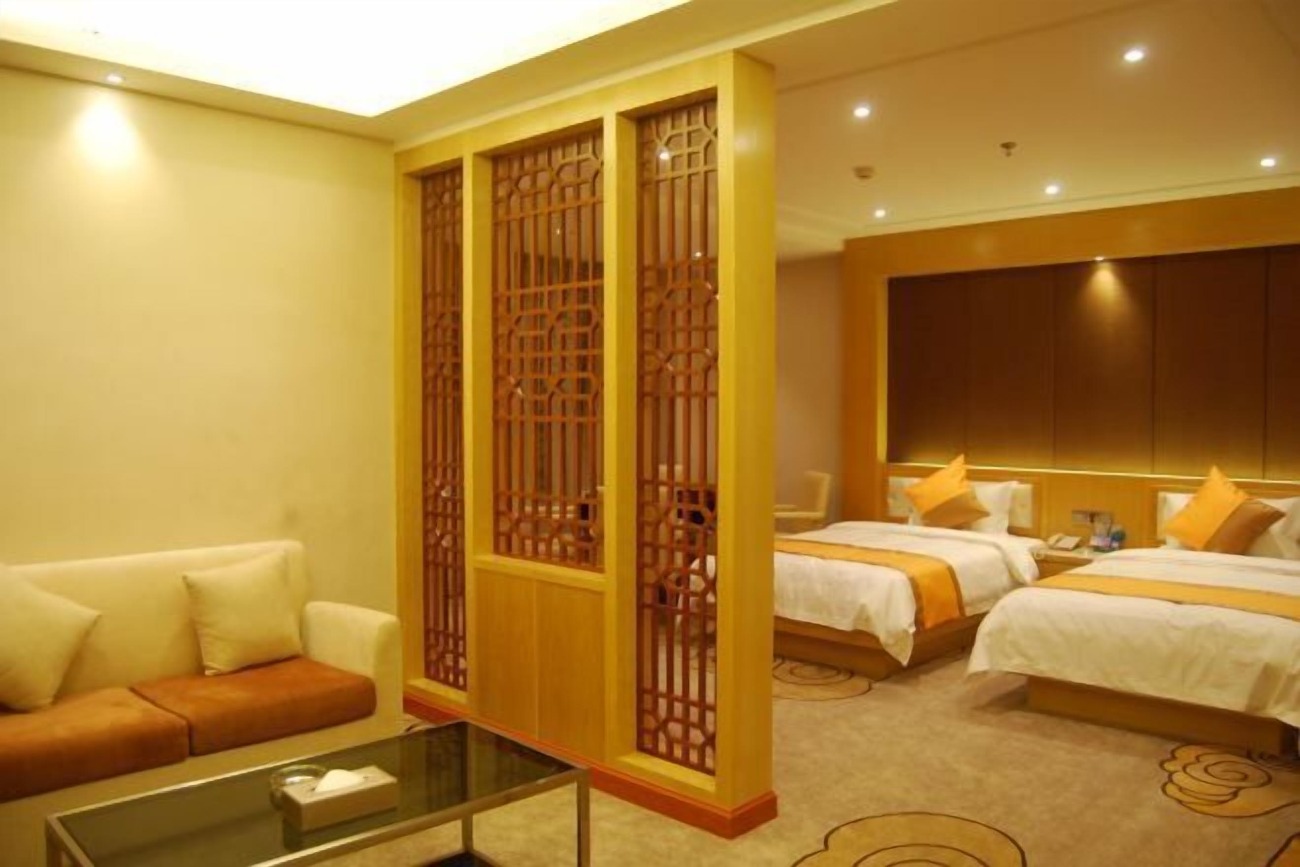 A hotel room with a sofa, two beds with white bedding and orange accents, a glass coffee table, and a decorative wooden screen.