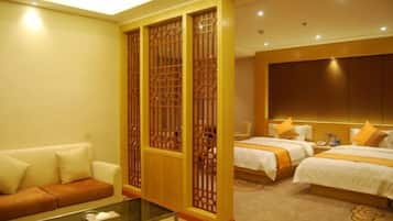 A hotel room with a sofa, two beds with white bedding and orange accents, a glass coffee table, and a decorative wooden screen.