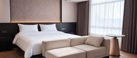 A modern hotel room with a large bed, a sofa, a small table, and a large window with curtains.