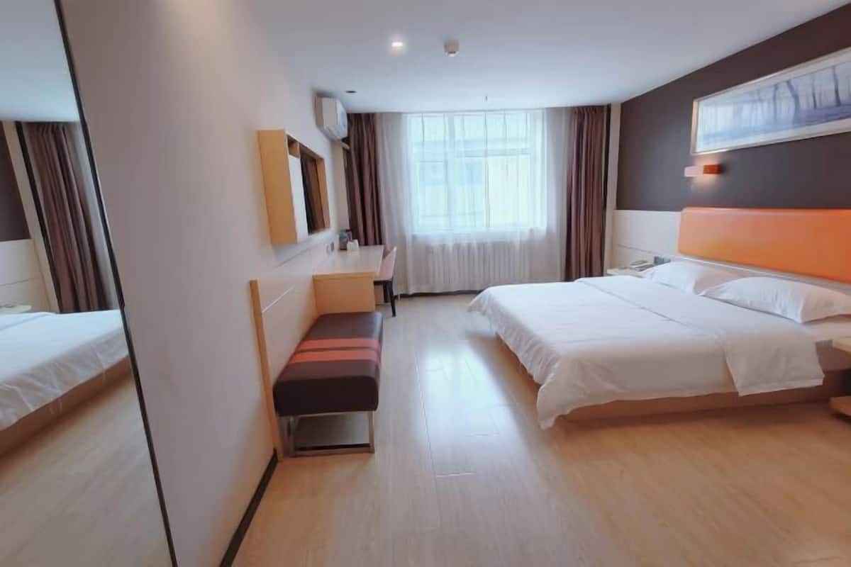 A hotel room with a bed, a bench, a desk, a chair, a window with curtains, and a painting on the wall.