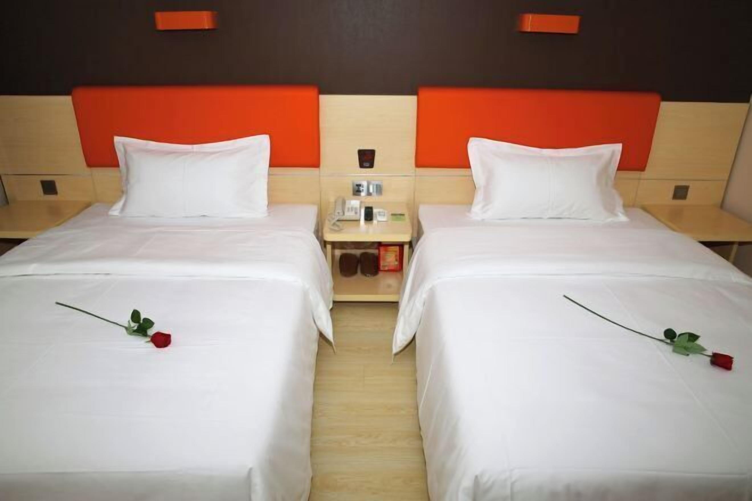 Habitación de hotel con dos camas, cada una con ropa blanca y una rosa roja. Entre las camas hay una mesita con un teléfono y una pequeña caja.