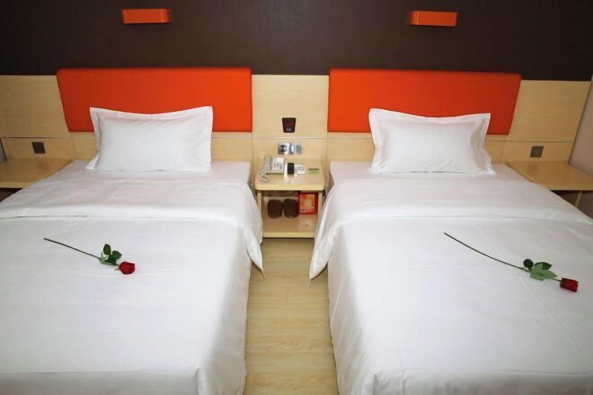 A hotel room with two beds, each with white bedding and a single red rose. There is a small table between the beds with a phone and a small box.
