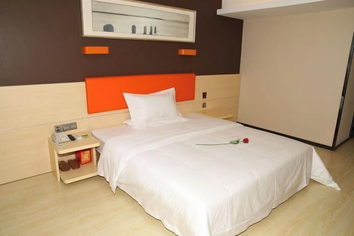 A hotel room with a bed, a nightstand with a phone and a box, a framed picture on the wall, and a door to the right.