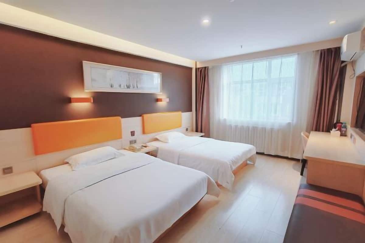 A hotel room with two beds, a desk, a chair, a window with curtains, and a wall-mounted artwork.